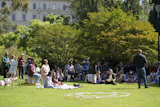 A group of people sit and stand on the grass in a park, listening to a speaker during an outdoor gathering, with trees and a historic building behind.